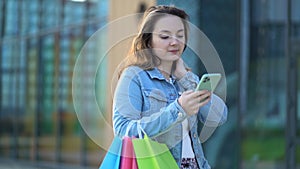 woman with multi-colored paper bags from the store walks down the street and uses her phone to communicate.