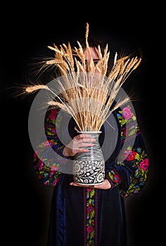 Woman in national dress holding a bouquet of dry wheat in a vase. On black bakground. Face covered with wheat