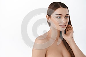 Woman with natural clean smooth skin, standing in profile, looking aside while touching straight brown healthy hair