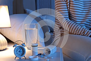 Woman near nightstand with pills, water and alarm clock in bedroom at night, closeup