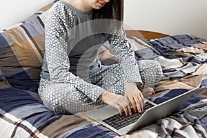 Woman in nightwear sits in the bed and uses her laptop. Home office during pandemic of coronavirus. Freelancer is typing.