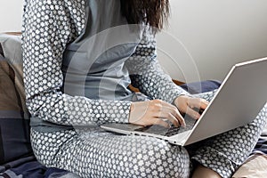 Woman in nightwear sits in the bed and uses her laptop. Home office during pandemic of coronavirus. Freelancer is typing using a