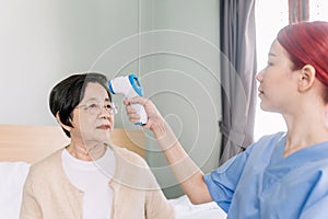 Woman nurse measures the temperature of an elderly Asian woman by using an infrared forehead thermometer gun at her home.