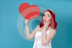 woman with open mouth in white dress and red hair holding a big red paper heart and holding her face