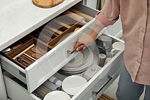 Woman opening drawers of kitchen cabinet with different dishware and utensils, closeup