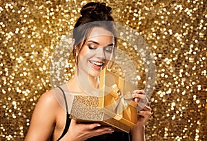 Woman opening Gift Box. Happy Girl holding Golden Present over Gold Glitter background. Beauty Model get Surprise on Christmas