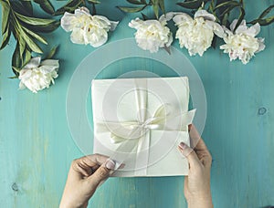 Woman opening her present, top view. Female`s hands pull ribbon to unwrap gift box among the white peony flowers on wooden