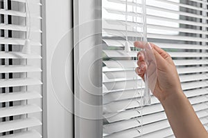 Woman opening horizontal blinds on window indoors, closeup. Space for text