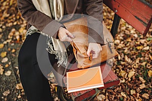 Woman opening leather handbag on bench in autumn park