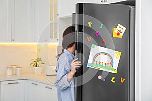 Woman opening refrigerator door with child`s drawing, notes and magnets