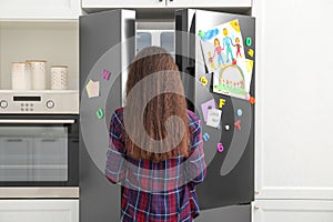 Woman opening refrigerator door with drawings, notes and magnets in kitchen