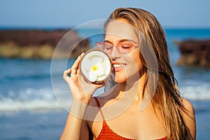 Woman in orange coral bikini and cool pink glasses standing on the beach with fresh dry open coconuts in hand. female