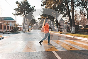 A woman in an orange jacket, walking alone on a pedestrian crossing. In the background, an empty road and street. In