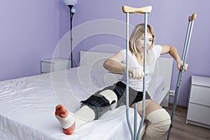 A woman in an orthosis and an elastic bandage after leg surgery sits on a bed in a hospital. Crutches in the postoperative period