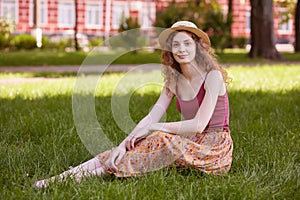 Woman outdoor portrait. Happy young woman sitting on green grass in park in summer, charming female wearing floor skirt, casual