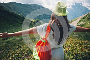 Woman with outspread hands standing among beautiful mountain meadows, wearing hat and backpack