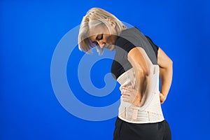 Woman in pain from back injury wearing lumbar brace corset on a blue background, copy space