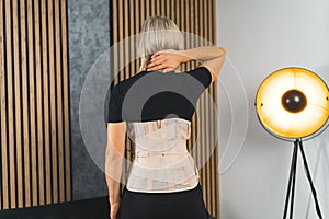 Woman in pain from back injury wearing lumbar brace corset at home, medium back view shot
