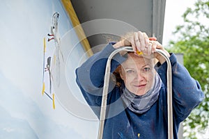 Woman, painter, artist in her forties or fifties with freckles and red hair stands on a ladder with a small brush in her hand,