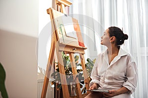 Woman painter in white shirt standing in front of easel thinking and looking at canvas