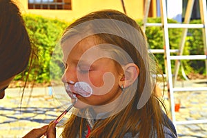 Woman painting face of kid outdoors. Baby face painting. Little girl getting her face painted like a rabbit by face painting artis