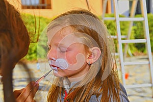 Woman painting face of kid outdoors. Baby face painting. Little girl getting her face painted like a rabbit by face painting artis