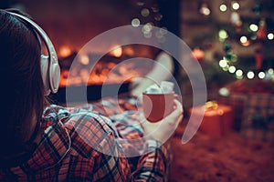 Woman in pajama in headphones with cup of hot cocoa and marshmallow sitting and warming at winter evening near fireplace flame.