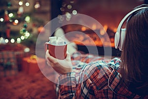 Woman in pajama in headphones with cup of hot cocoa and marshmallow sitting and warming at winter evening near fireplace flame.