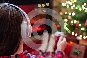 Woman in pajama in headphones with cup of hot cocoa and marshmallow sitting and warming at winter evening near fireplace flame.