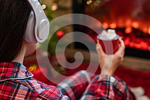 Woman in pajama in headphones with cup of hot cocoa and marshmallow sitting and warming at winter evening near fireplace flame.