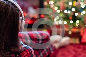 Woman in pajama in headphones sitting and warming at winter evening near fireplace flame and  christmas tree.