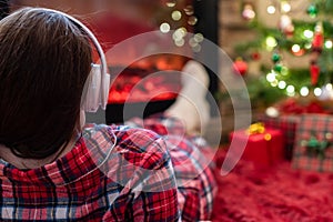 Woman in pajama in headphones sitting and warming at winter evening near fireplace flame and  christmas tree.