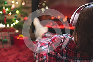 Woman in pajama in headphones sitting and warming at winter evening near fireplace flame and  christmas tree.