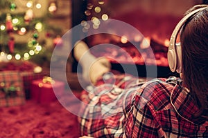 Woman in pajama in headphones sitting and warming at winter evening near fireplace flame and  christmas tree.