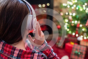 Woman in pajama in headphones sitting and warming at winter evening near fireplace flame and  christmas tree.