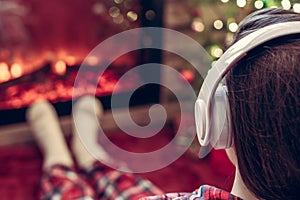 Woman in pajama in headphones sitting and warming at winter evening near fireplace flame and  christmas tree.