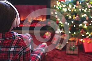Woman in pajama in headphones sitting and warming at winter evening near fireplace flame and  christmas tree.
