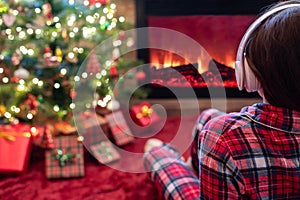 Woman in pajama in headphones sitting and warming at winter evening near fireplace flame and  christmas tree.