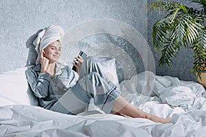 A woman in pajamas and with a towel on her head sits on a bed in the morning sun and listens to music on headphones through the