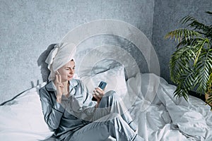 A woman in pajamas and with a towel on her head sits on a bed in the morning sun and listens to music on headphones through the
