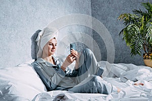 A woman in pajamas and a towel on her head sits on a bed in the morning sun and looks at something on her phone