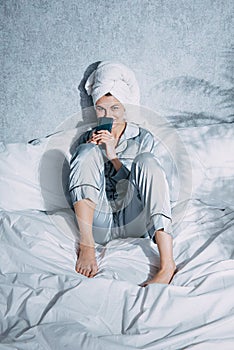 A woman in pajamas and a towel on her head sits on a bed in the morning sun and looks at something on her phone