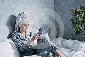 A woman in pajamas and a towel on her head sits on a bed in the morning sun and makes online purchases using her mobile phone
