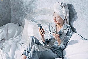 A woman in pajamas and a towel on her head sits on a bed in the morning sun and makes online purchases using her mobile phone