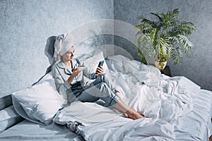A woman in pajamas and a towel on her head sits on a bed in the morning sun and makes online purchases using her mobile phone