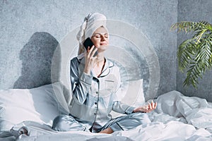 A woman in pajamas and with a towel on her head sits on a bed in the morning sun and meditates talking on the phone