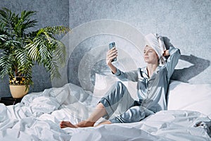 Woman in pajamas and a towel on her head sits on a bed in the morning sun and takes a selfie