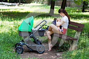 Woman parent is sitting on wooden bench in the park, holding and breastfeeding baby, next to stroller, sunny day in public park