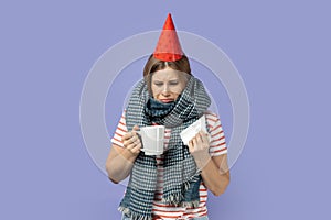 Woman in party cone, female celebrating her birthday and feels unwell holding cup with tea and