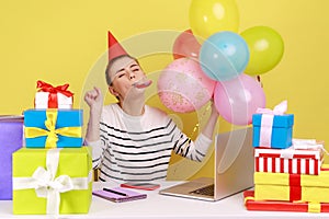 Woman with party cone sitting workplace among gifts and balloons, blowing horn, celebrating birthday